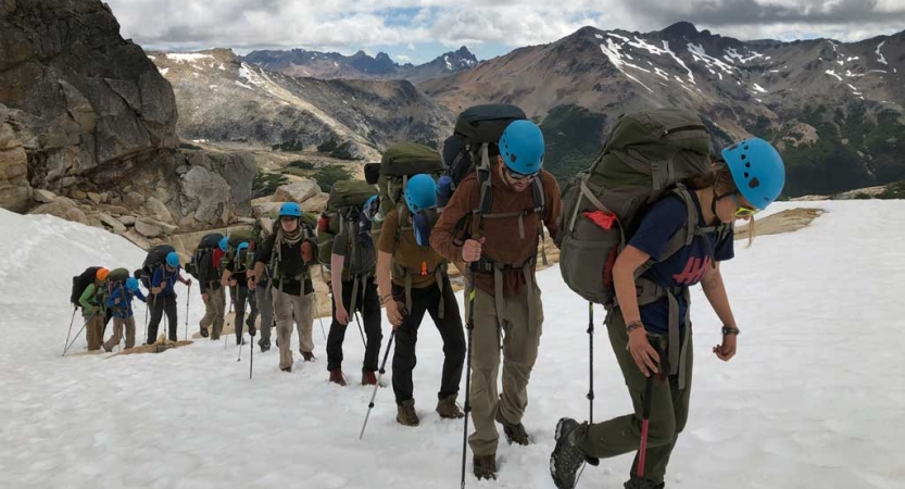 A group of people carrying backpacks and trekking poles, and wearing helmets, hike in a line up a snowy incline. Behind them is a vast mountainous landscape.